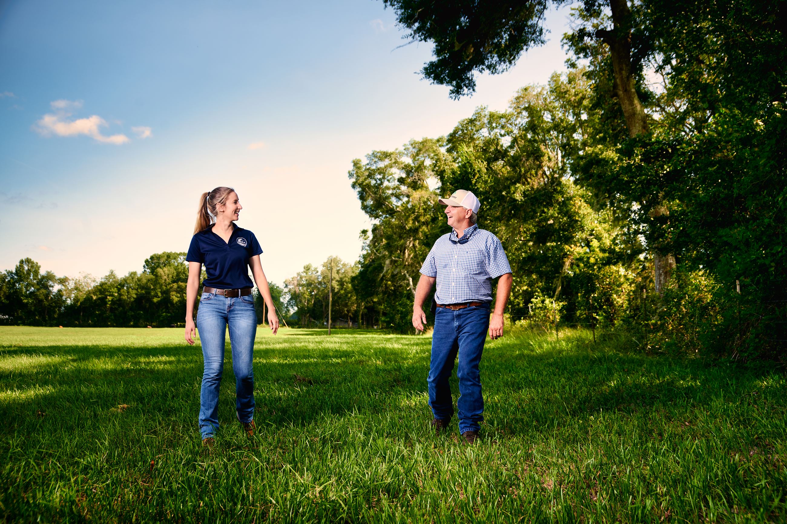 Two people walking in a pasture lined with trees.